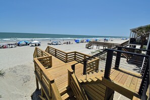 Eine Holzterrasse mit Geländer, die zu einem Strandbereich mit Sonnenschirmen und Liegestühlen führt.