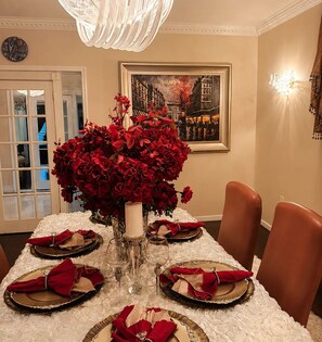 Una mesa de comedor preparada para seis personas, con servilletas rojas, un florero con flores rojas y un cuadro en la pared.