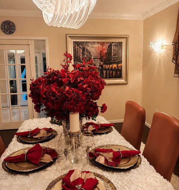A dining table set for six with red napkins, a vase of red flowers, and a painting on the wall.