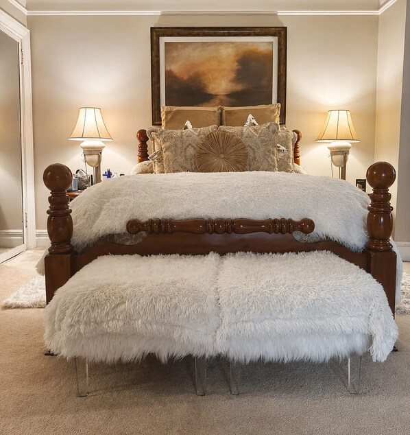 Un dormitorio con una cama grande, dos lámparas de noche, una obra de arte enmarcada sobre la cama y un banco blanco mullido frente a la cama.