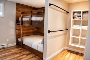 A room with a bunk bed, a white shelving unit with folded towels, and a wooden floor.