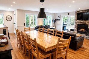 Ein geräumiges Wohnzimmer mit einem großen Esstisch aus Holz, bequemen Sitzgelegenheiten, einem Kamin und einem an einer Steinwand befestigten Fernseher.