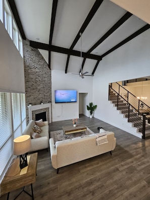 A spacious living area with a fireplace, a flat-screen TV, a staircase, and a large window. - Songbird Luxury Retreat — a Modern 4BR Getaway in Wylie, Near Lake Lavon (Wylie)