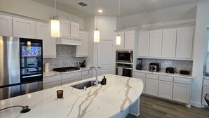 Une cuisine moderne avec des meubles blancs, un grand îlot central doté d’un évier, des appareils en acier inoxydable, des luminaires suspendus et un plancher en bois.