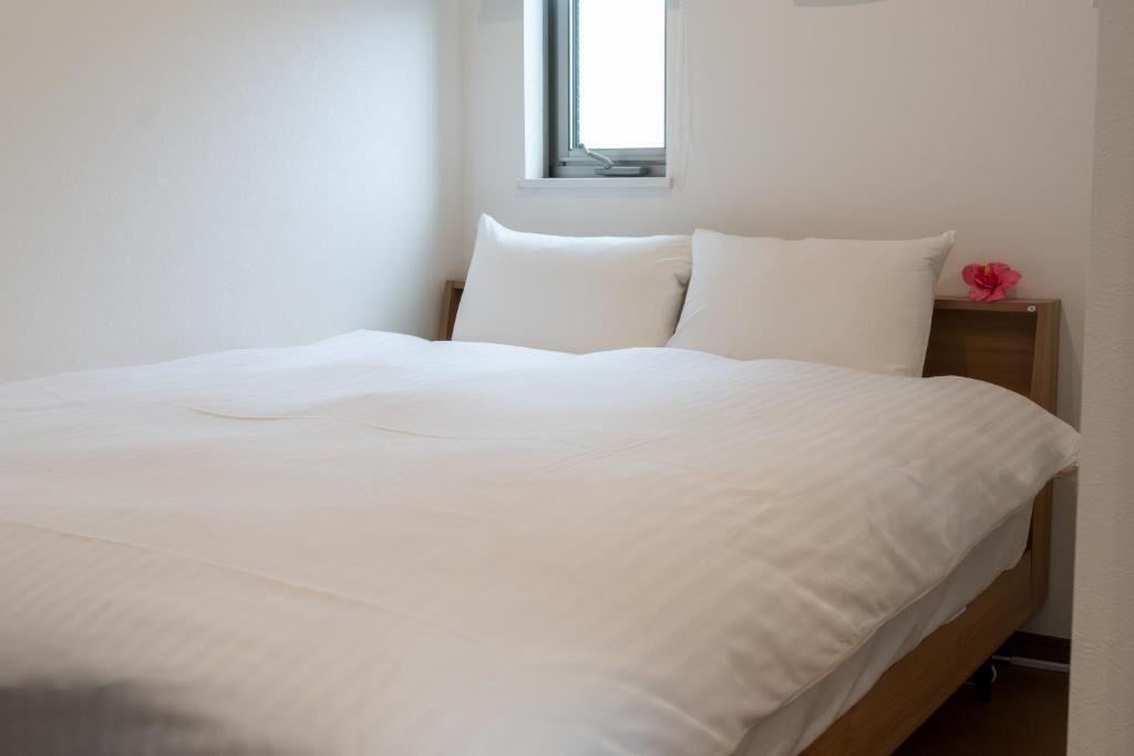 A bed with white bedding in a room with a window and a nightstand with a pink flower.
