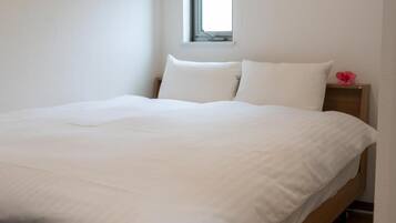 A bed with white bedding in a room with a window and a nightstand with a pink flower.
