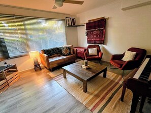 A living room with a sofa, armchairs, a coffee table, a piano, and a window with blinds.