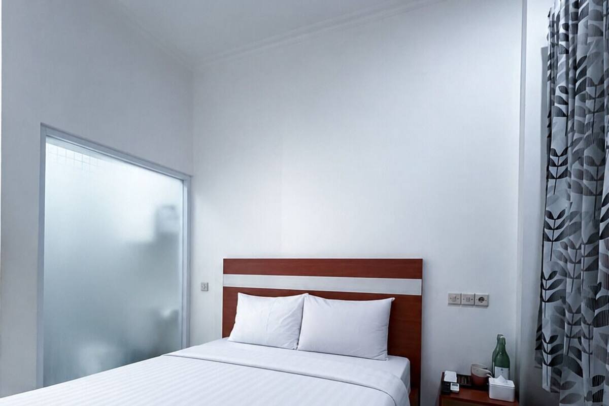A hotel room with a bed, a nightstand, a lamp, a window with a view, and a door.