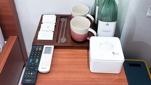 Une table en bois avec une télécommande, deux tasses, une bouteille, un récipient blanc et un plateau avec des serviettes et une cuillère.