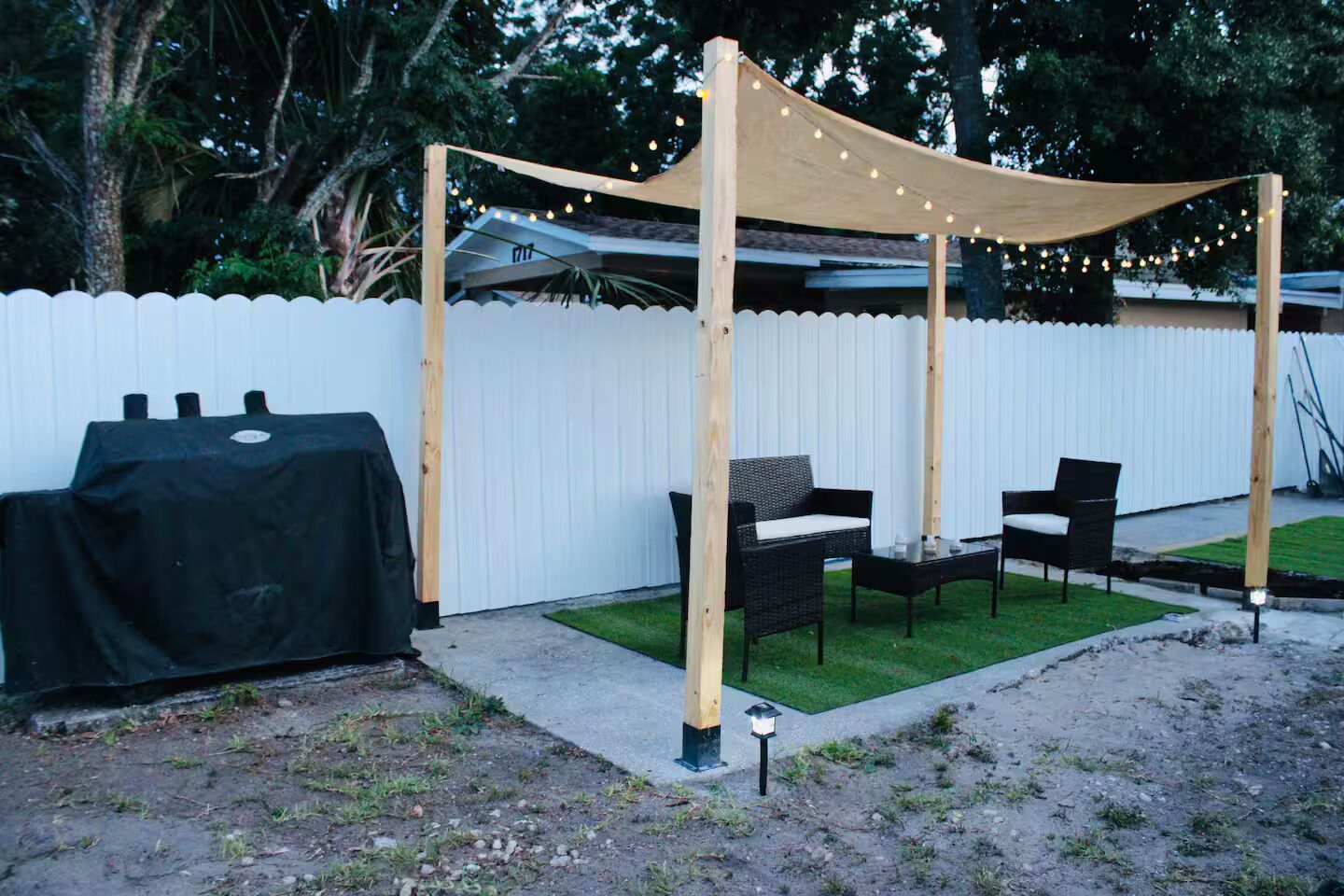 Un espace extérieur aménagé avec une zone de repos couverte, comprenant des chaises et une table, un barbecue et, au fond, une clôture blanche.