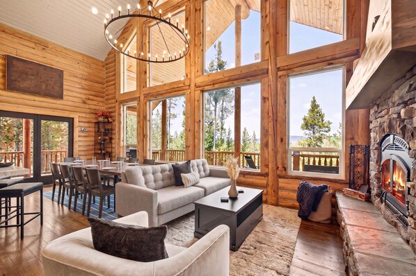 L’intérieur d’une cabane en rondins, avec une cheminée, de grandes fenêtres, un coin repas et un espace salon confortable.