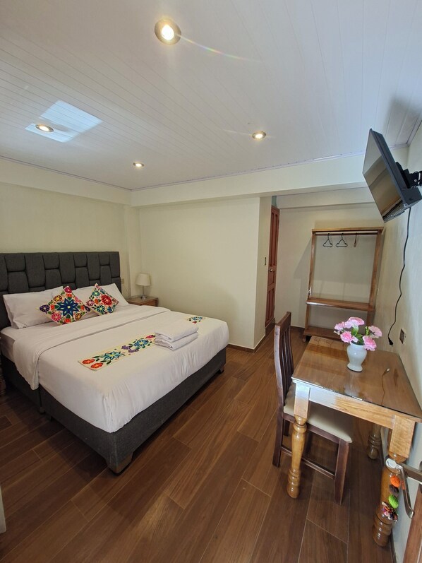 A bedroom with a bed, bedside tables, a desk, a chair, a TV mounted on the wall, and a wooden floor.