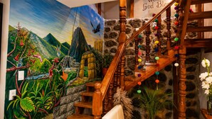 A wooden staircase with colorful pom-poms, a mural with a bird and mountains, and a 'Welcome God Bless' sign. - HOTEL PIEDRA BLANCA (Ollantaytambo)