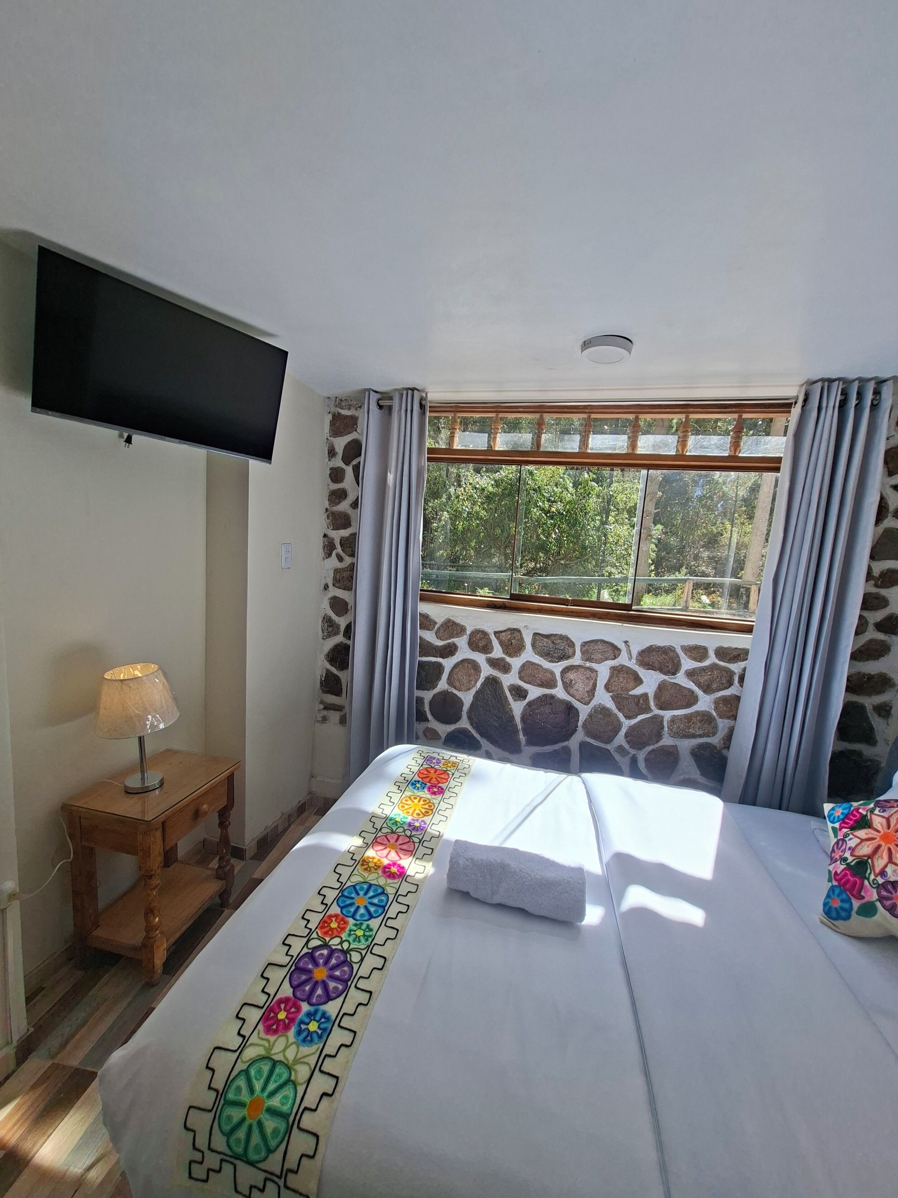 A hotel room with a bed, a TV, a lamp, a window with curtains, and a stone wall.