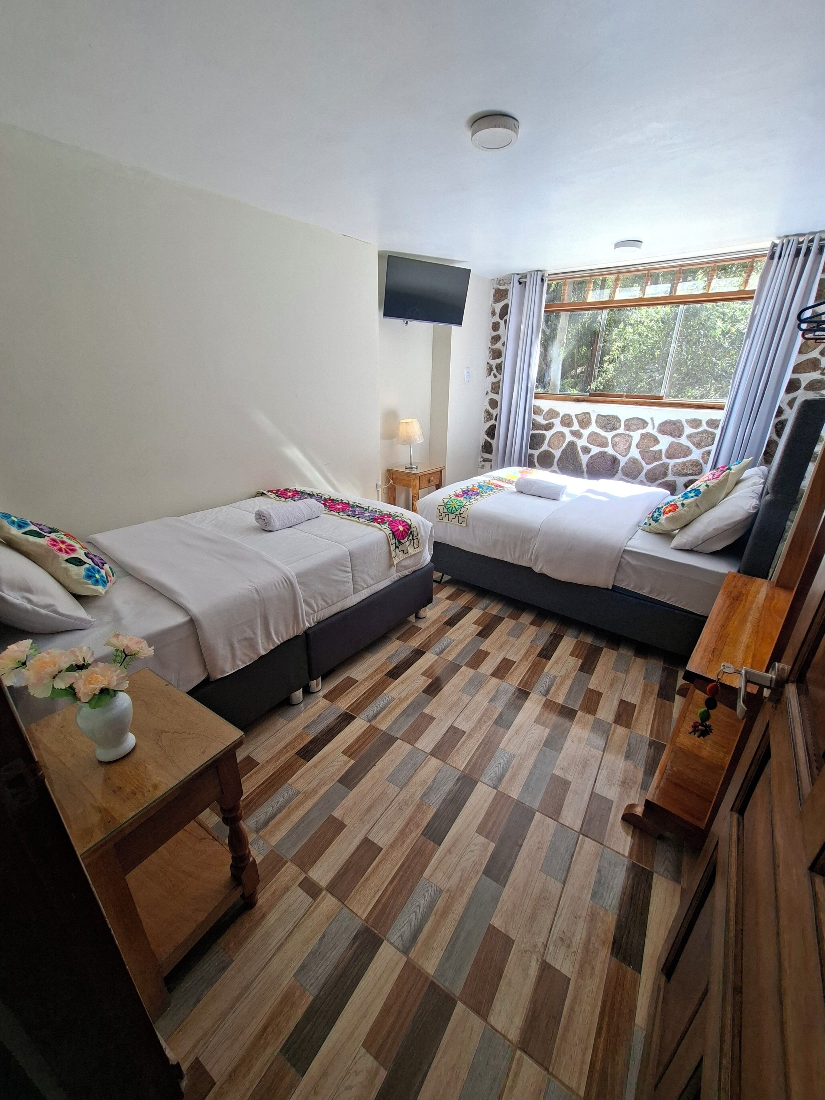 A room with two beds, a TV, a stone wall, and a window with curtains.