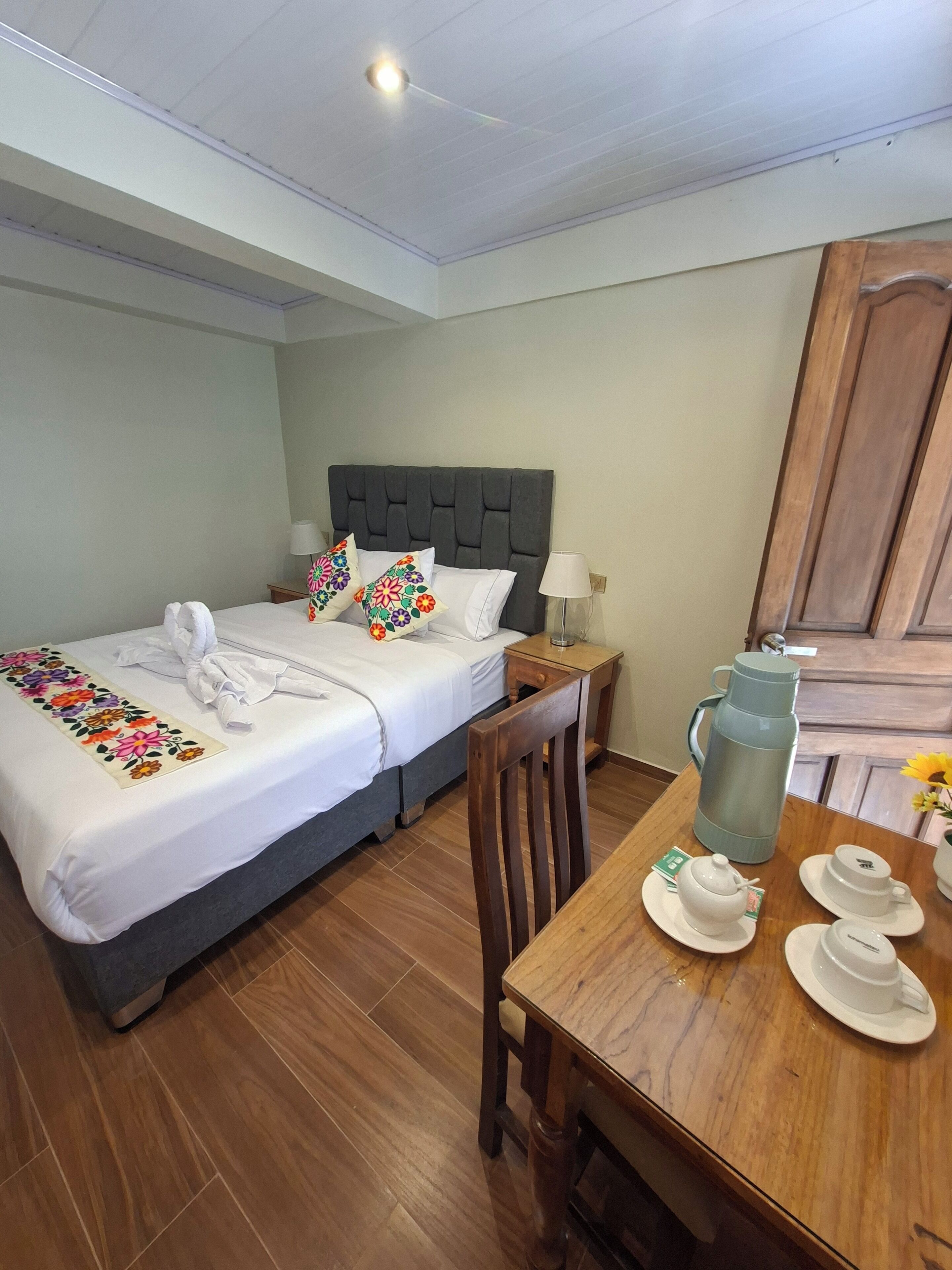Una habitación de hotel con una cama, una mesa de madera con juegos de té, un hervidor y una mesita de noche con una lámpara.