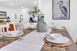 A dining area with a long table set for four, surrounded by chairs, with a kitchen in the background. - Remodeled Coastal Retreat with Breathtaking Views (North Myrtle Beach)