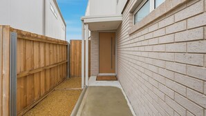 Un estrecho pasillo exterior con una cerca de madera a un lado y un muro de ladrillo al otro, que conduce a una puerta.