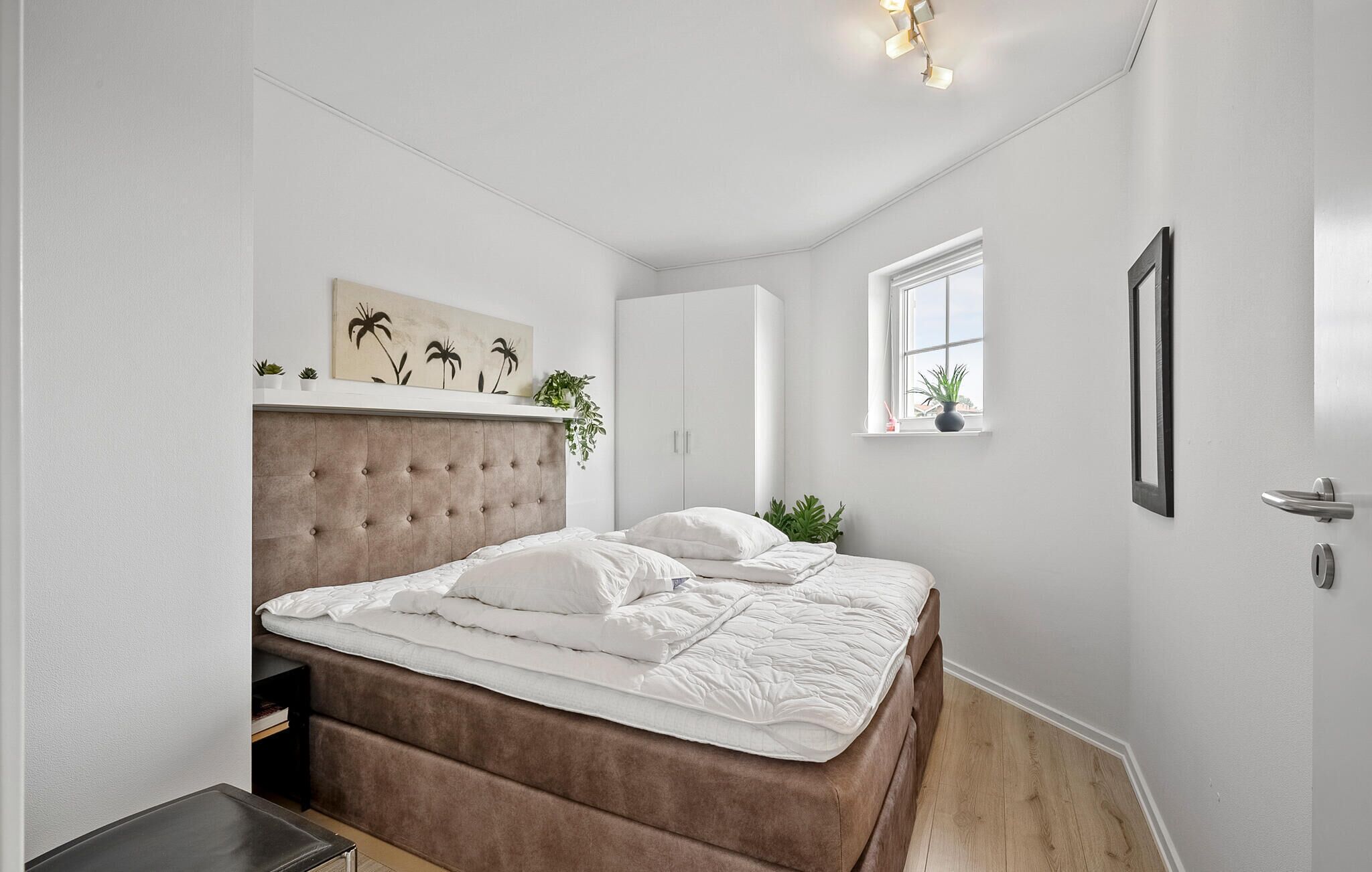 Un dormitorio con cabecera capitonada, cama con ropa de cama blanca, ventana con vistas y una planta en el alféizar.
