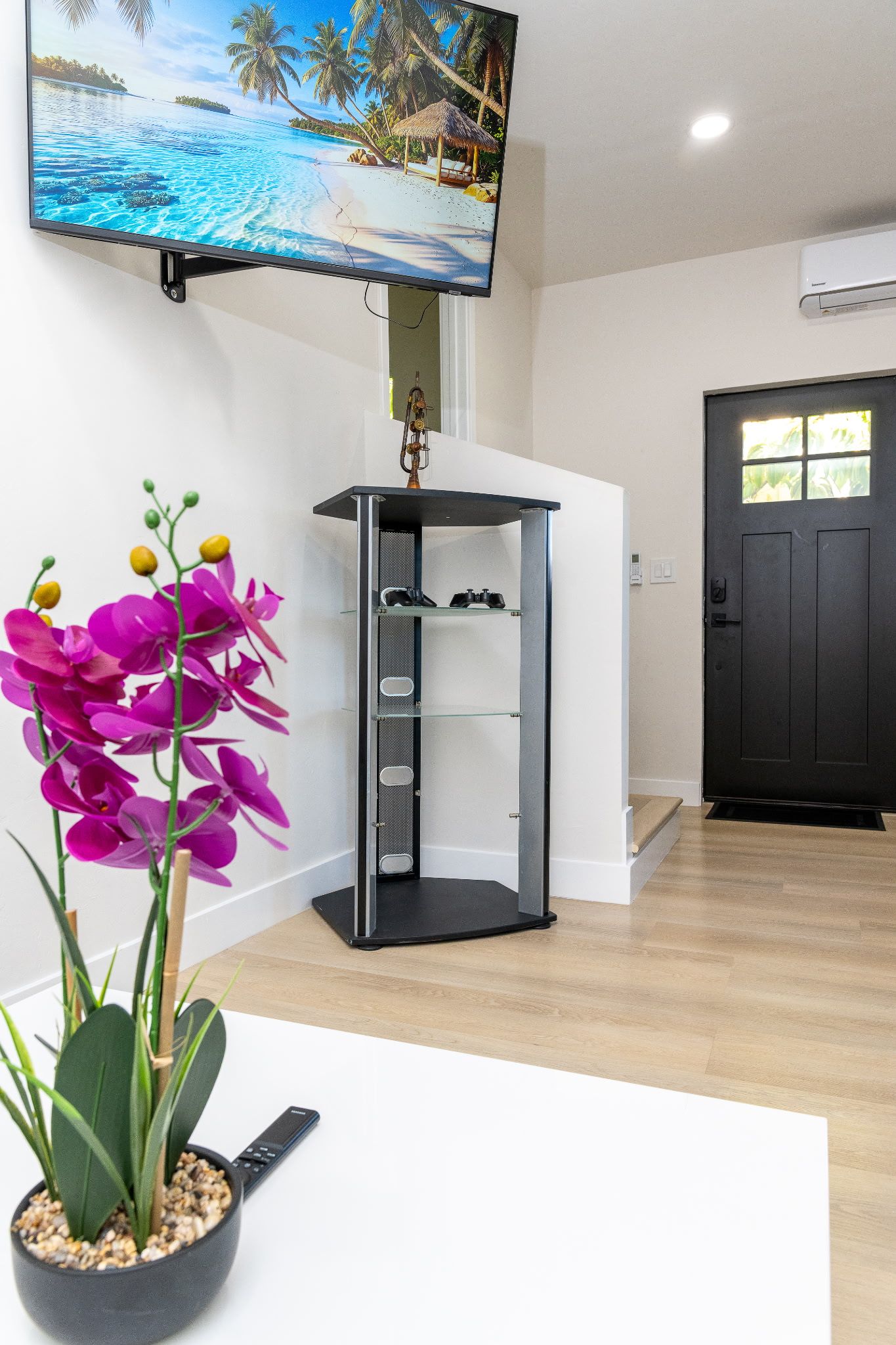 Una habitación con un televisor montado en la pared, una puerta negra y una planta con flores moradas sobre una mesa blanca.
