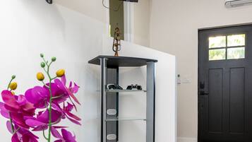 Una habitación con un televisor montado en la pared, una puerta negra y una planta con flores moradas sobre una mesa blanca.