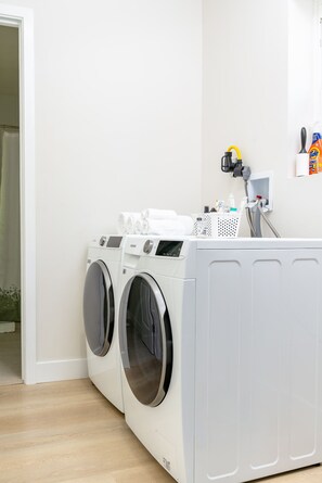 Un espace buanderie comprenant deux machines à laver à chargement frontal, un étendoir avec des serviettes pliées, et un meuble de rangement mural avec un tuyau et du détergent.