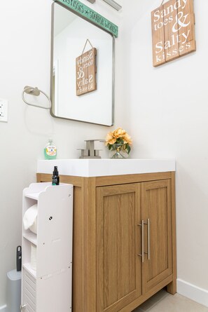 Une salle de bain avec un meuble en bois, un lavabo blanc et un miroir. Il y a deux panneaux accrochés au mur, l’un avec un message et l’autre avec un nom.