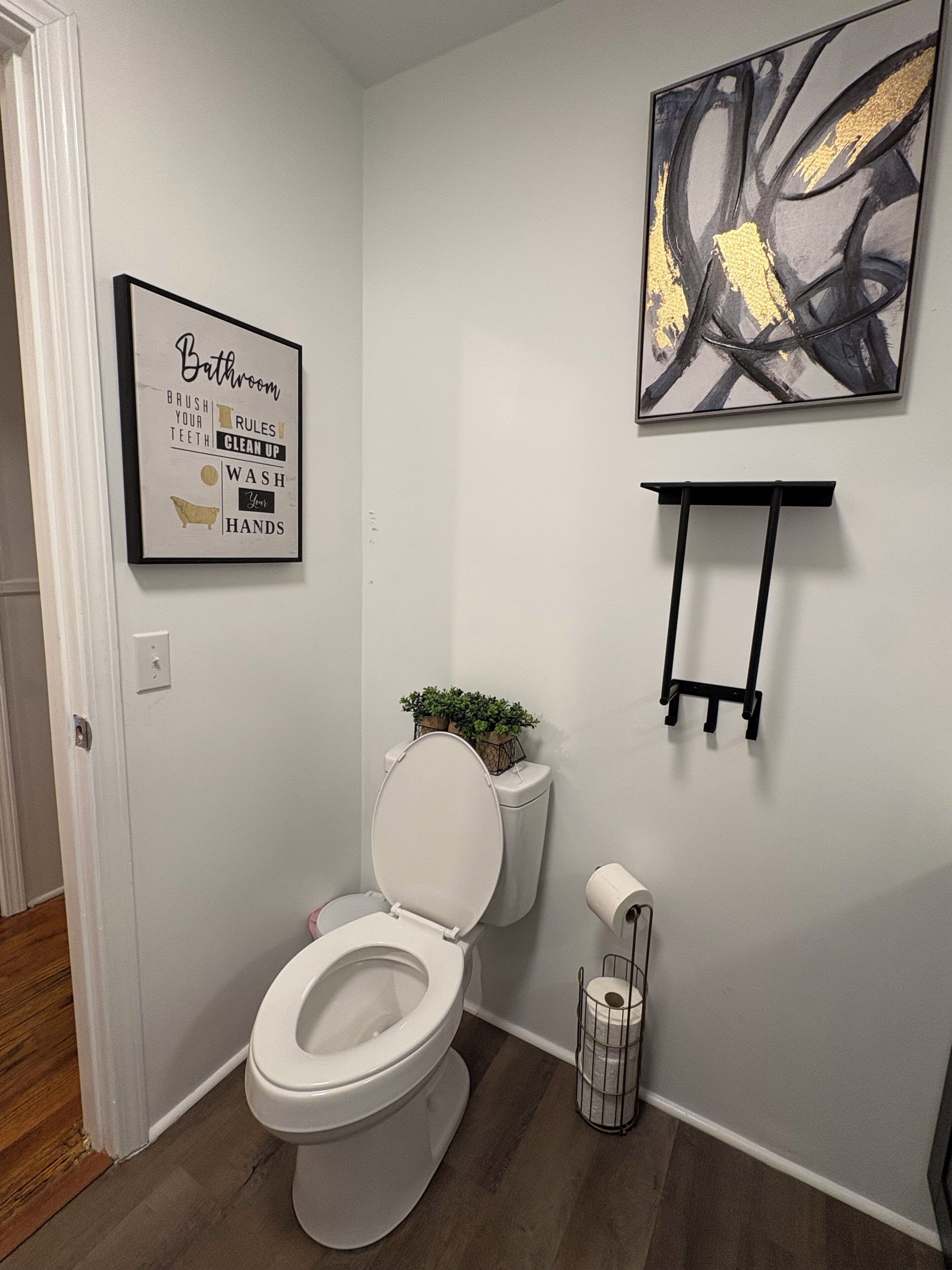 A bathroom with a toilet, a framed sign on the wall, a hanging abstract painting, and a small shelf.