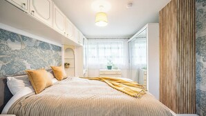 Een slaapkamer met een bed, nachtkastje, dressoir, raam met zonwering en een wand met een verticale houten lambrisering.