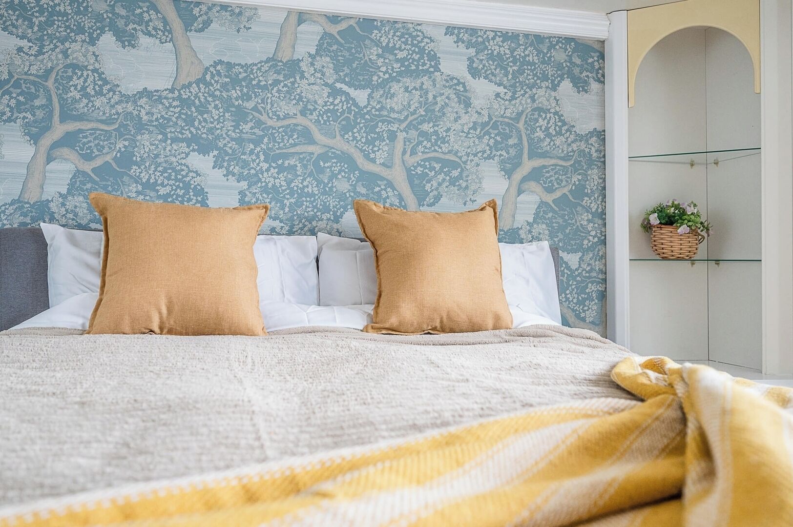 Un letto rifatto con una coperta a righe, una parete con un motivo alberato e un piccolo ripiano con una pianta.