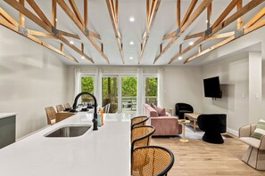 Eine moderne Küche mit weißer Arbeitsplatte und Holzbalken, ein Essbereich mit Korbstühlen und ein Wohnbereich mit Sofa und Fernseher.