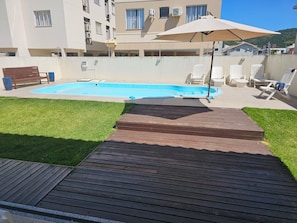 Una piscina con cubierta de madera, rodeada de césped y sillas blancas.