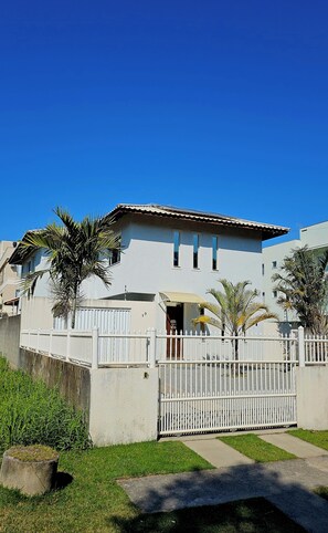 Un edificio de dos pisos con fachada blanca, entrada con reja y un pequeño jardín con palmeras.