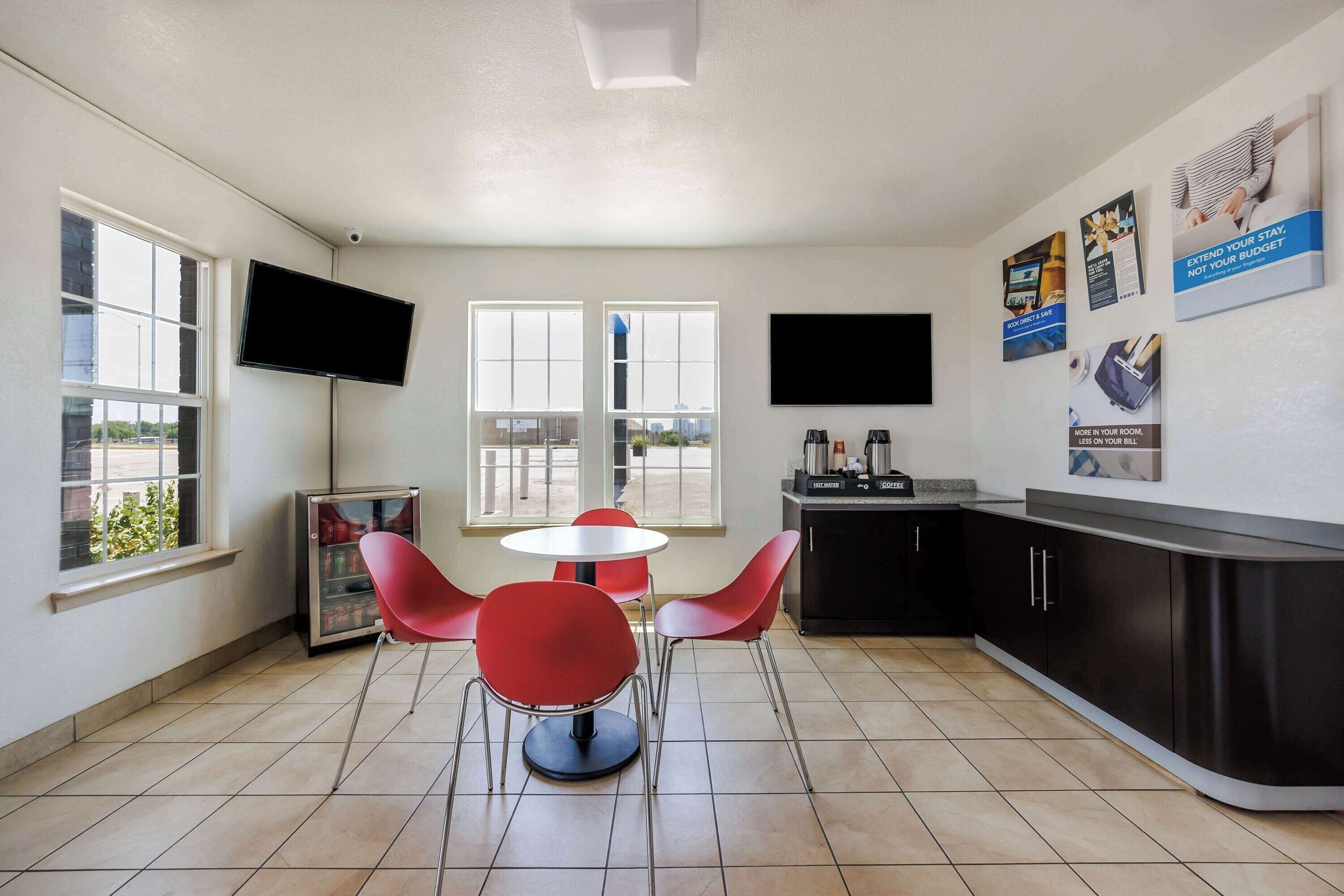 A modern kitchen with a refrigerator, microwave, and coffee maker, a dining area with red chairs and a small table, and a TV mounted on the wall.