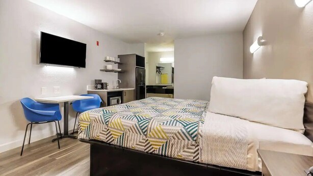 Habitación de hotel pequeña con cama, escritorio con sillas, televisor de pantalla plana y una kitchenette al fondo.