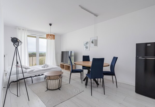 Quarto moderno com uma cama, uma mesa de jantar com cadeiras, um refrigerador preto e uma televisão sobre um suporte.