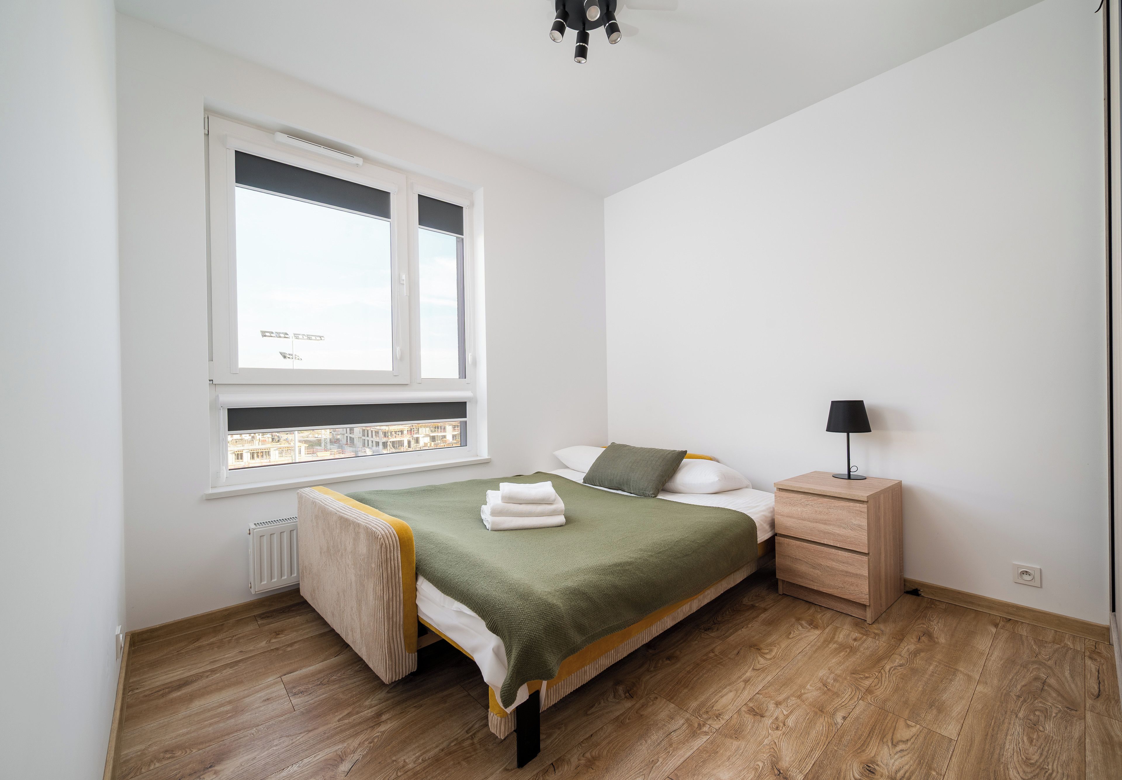 Un dormitorio con cama, mesita de noche, lámpara y ventana con vista a edificios.
