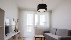 A modern living room with a grey sofa, a small table, a TV on a white shelf, and a ceiling light. - Długa Homely Apartment (Toruń)