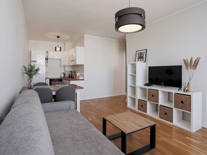 Un salon moderne avec un canapé, une table basse, un téléviseur posé sur un meuble blanc, et une cuisine en arrière-plan.