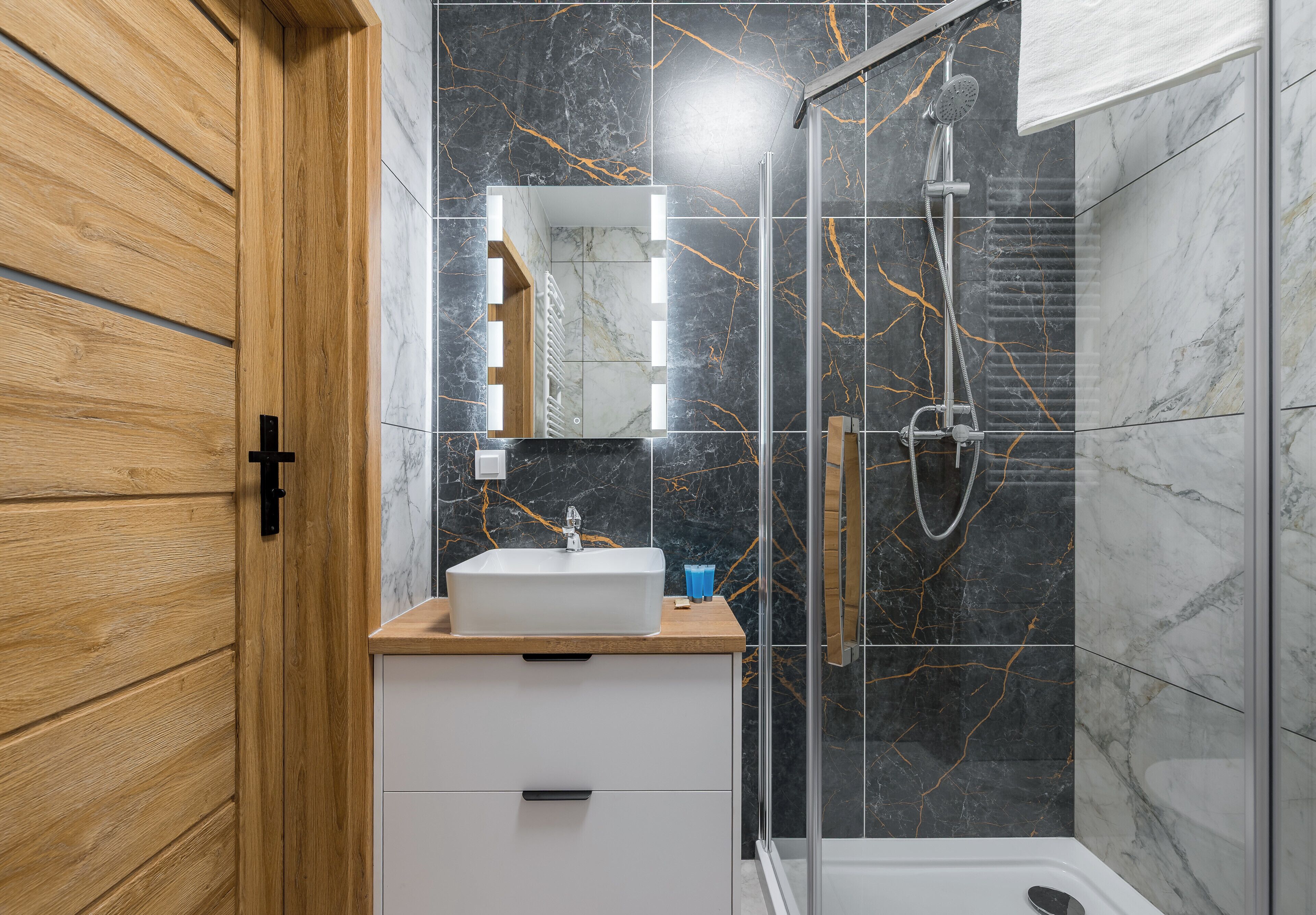 Une salle de bain moderne avec une porte en bois, un lavabo blanc, un miroir avec éclairage intégré, une douche et des murs en marbre.