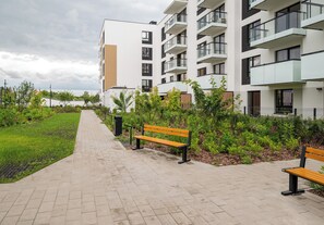 Ein gepflasterter Weg mit Bänken, umgeben von Grünflächen und einem modernen Wohnhaus.
