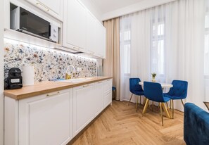 Eine moderne Küche mit weißen Schränken, einer Mikrowelle und einer Rückwand mit floralem Muster. Es gibt eine Essecke mit einem runden Tisch und blauen Stühlen.