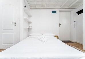 Ein heller Raum mit einem Bett, einer Tür, einem Regal mit einer kleinen Pflanze und einem Thermostat an der Wand.