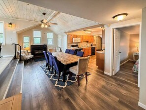 A spacious living area with a dining table, sofas, and a fireplace. There is a kitchen in the background with wooden cabinets and a microwave.