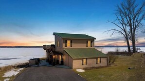 A two-story house with a green roof, a front porch, and a view of a body of water. - Lower Lake Retreat | Charming Waterfront Stay (Seneca Falls)