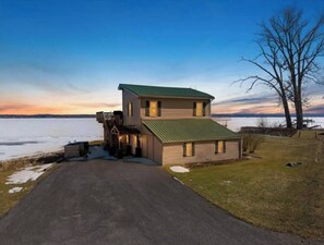 A two-story house with a green roof, a front porch, and a view of a body of water.