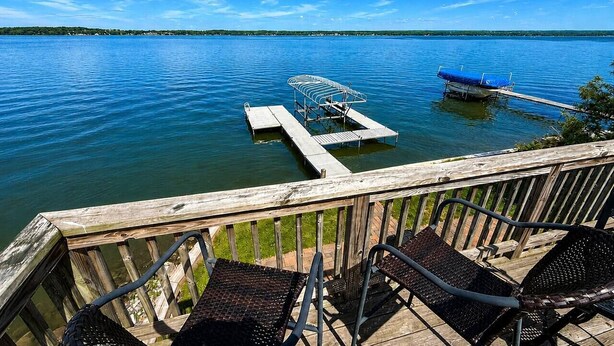 A wooden deck with metal chairs overlooking a calm lake, a dock with a boat, and a distant shoreline. - Lower Lake Retreat | Charming Waterfront Stay (Seneca Falls)