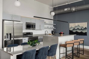 A modern kitchen with stainless steel appliances, a central island with bar stools, and a hanging light fixture. - The Ridgeline at Murals on Market (Chattanooga)
