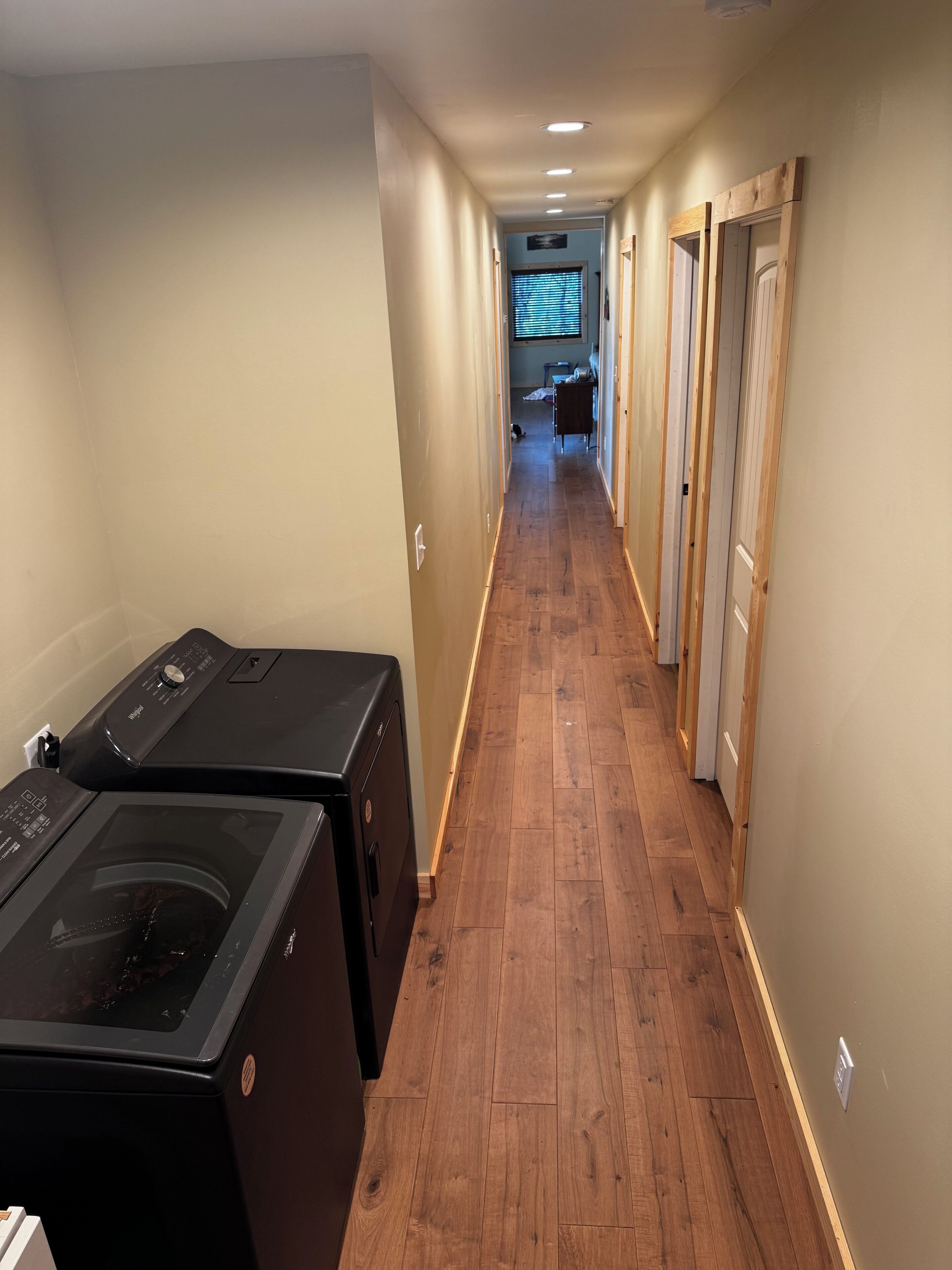 A laundry room with washers and dryers, a wooden floor, and a hallway leading to a room with a window.