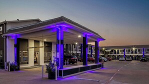 A motel with a covered parking area, multiple cars parked, and a clear sky. - 2 Vibrant 2-Bed Studio | Kitchenette, Free Parking | Easy Highway Access (Fort Worth)